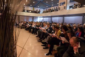 In een gevulde zaal zitten mensen met naamkaartjes op hun borst. Iedereen heeft een papier vast en kijkt daarnaar.