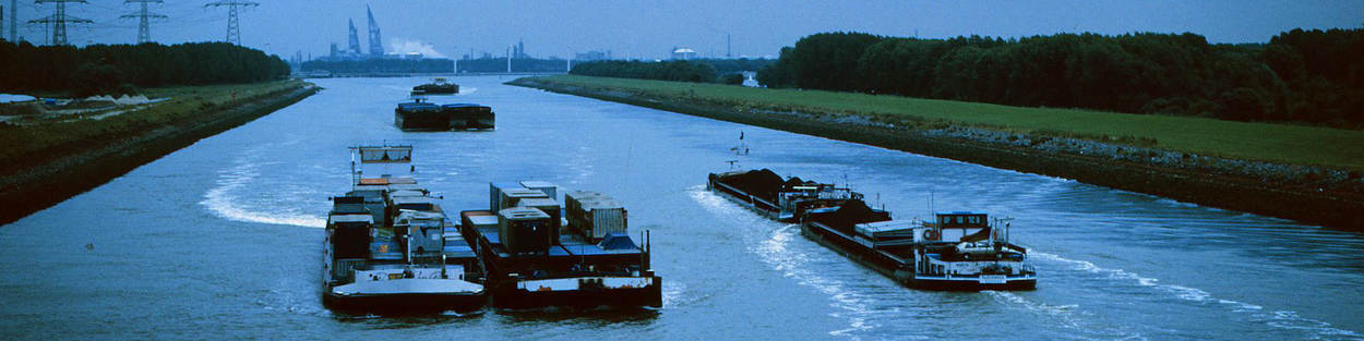 Een aantal binnenvaartschepen varen naast elkaar over een kanaal. Op de achtergrond zie je een industriegebied.