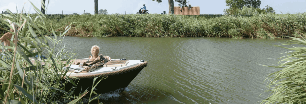 Een gemotoriseerd bootje vaart in de zon door een kanaal. Op het bootje zitten een man en een vrouw.