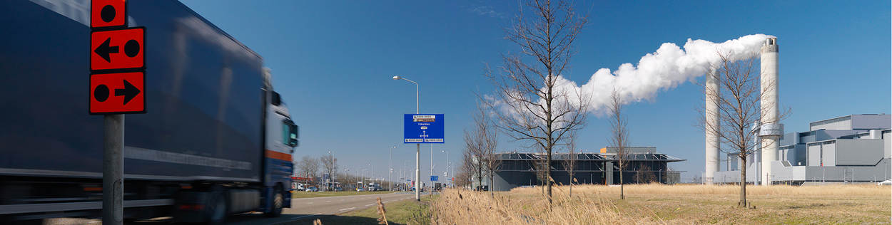 Een vrachtwagen rijdt over een weg naar een industriegebied. Op de voorgrond staan bordjes die de routes voor transport met gevaarlijke stoffen aanduidt. Op de achtergrond staat een fabriek met flinke rookpluimen.