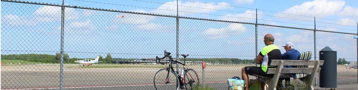 Twee vliegtuigspotters zitten op een bankje bij de start- en landingsbaan van vliegveld Lelystad. Op de achtergrond staat een klein sportvliegtuig.