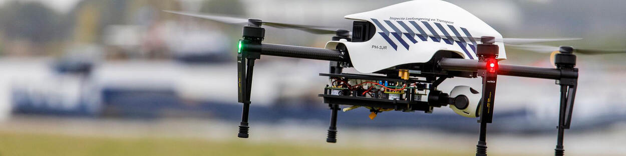 Een drone van de Inspectie Leefomgeving en Transport hangt in de lucht. Hij is herkenbaar als inspectiedrone door de blauwe striping op de witte buitenkant. Achter de drone vaart een binnenvaartschip.
