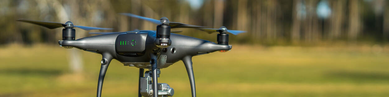 Een grijze drone met vier wieken hangt in de lucht. Op de achtergrond is een weiland met daarachter een bosrand.