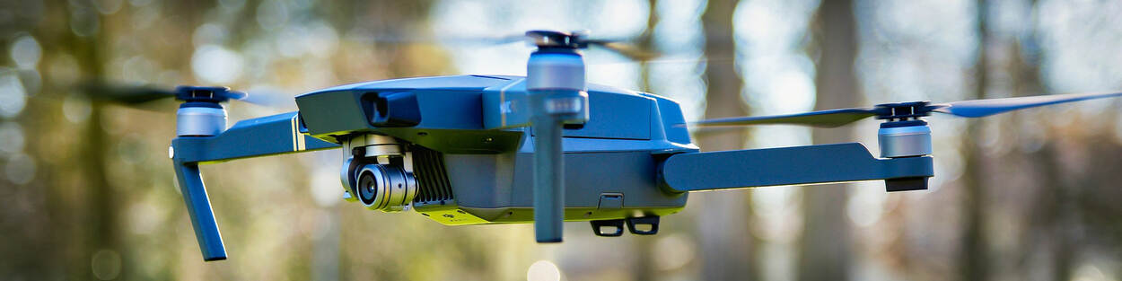 Een blauwgekleurde drone hangt in de lucht. Hij heeft een camera en vier wieken. Op de achtergrond zijn de contouren van enkele bomen.