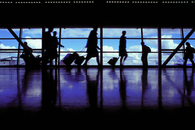 Passagiers lopen met koffers door luchthaven Schiphol. Door het glas is op de achtergrond een vliegtuig zichtbaar.