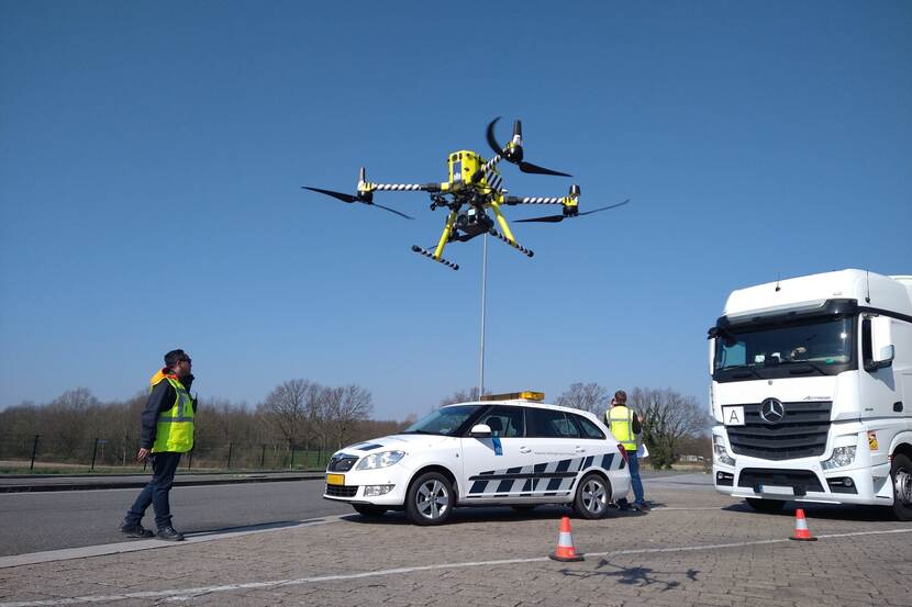 Parkeerterrein met aangehouden vrachtwagen met afvaltransport. Twee inspecteurs bij hun dienstauto lanceren een drone die het afvaltransport van bovenaf gaat bekijken.