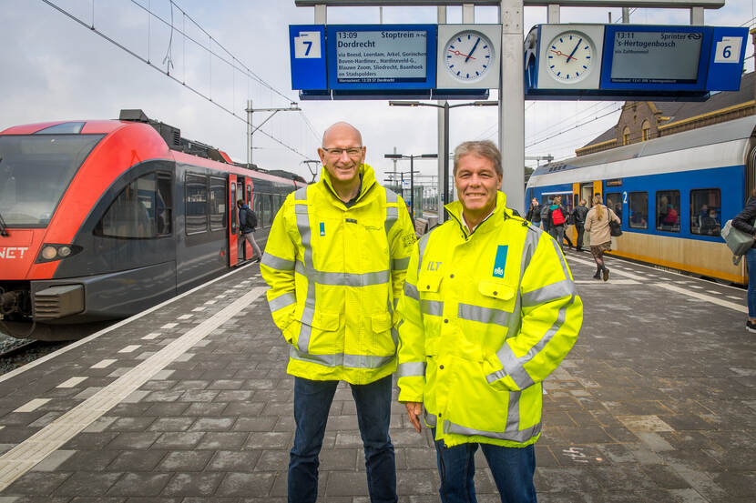 Gert van der Rijst en Adri Legierse staan samen op een perron in ILT-jassen. Op de achtergrond staan twee treinen.