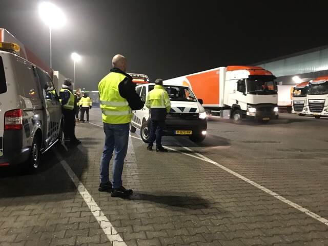 Enkele inspecteurs staan in het donker op een parkeerplaats en controleren vrachtwagens.
