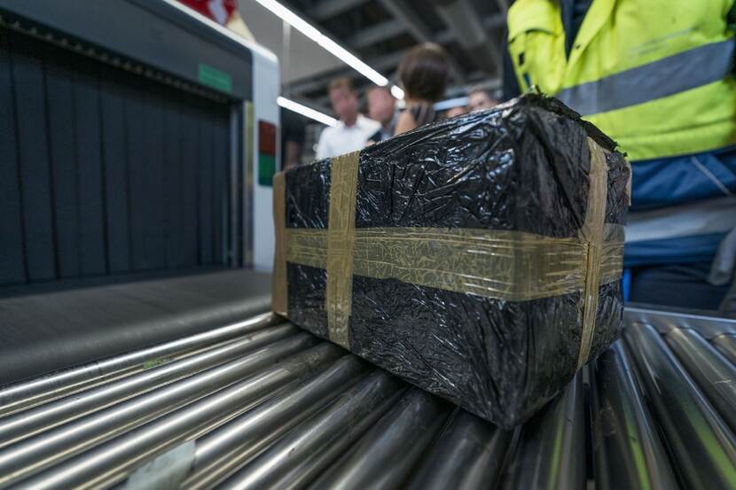 Een postpakket staat op een lopende band nabij een scanapparaat. Op de achtergrond staat een inspecteur.