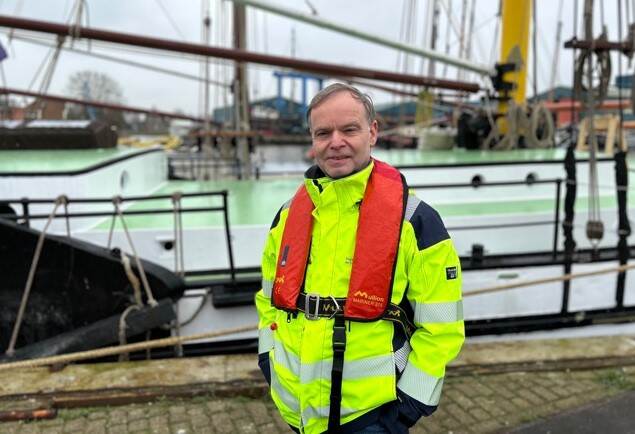 Een man in een fluorgele veiligheidsjas staat voor een historisch zeilschip.