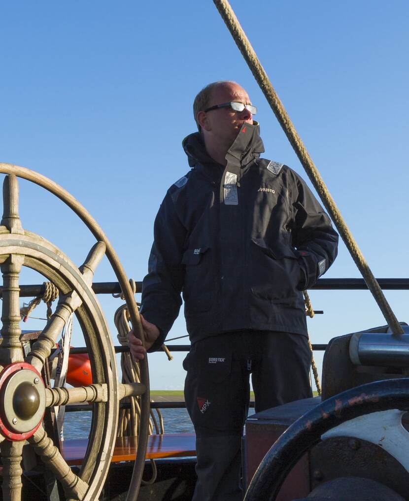 Een schipper in een windjack draait aan een groot houten stuurwiel op een historisch zeilschip