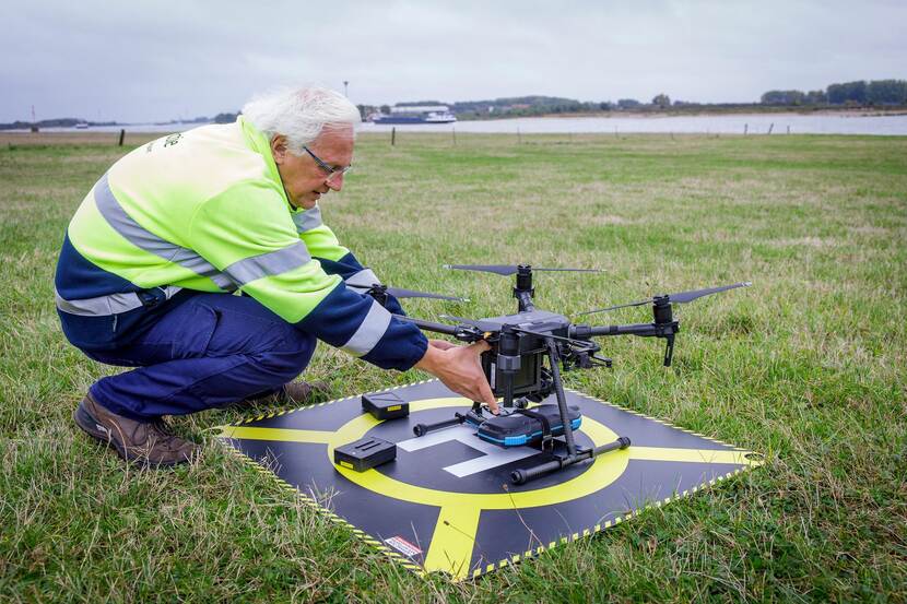 Een inspecteur zit op zijn hurken naast een opstijgplatform voor drones. Op het platform staat een ILT-drone. Op de achtergrond vaart een schip voorbij.