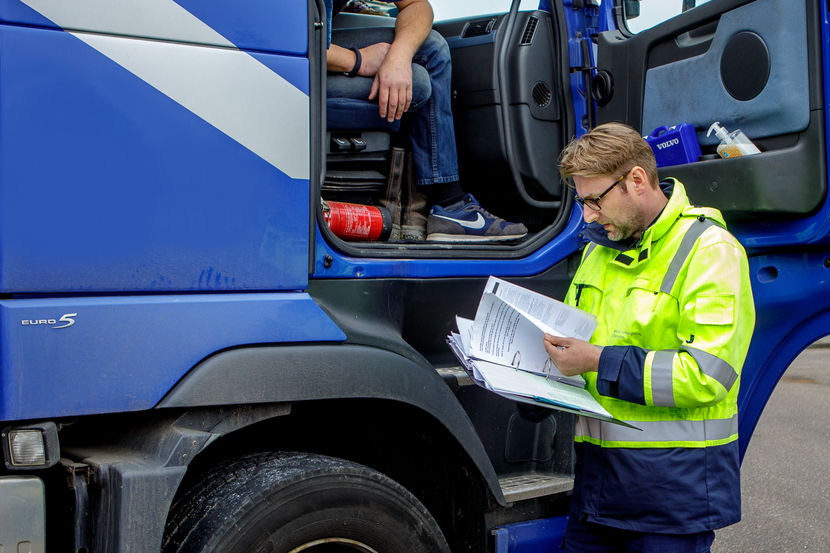 Een inspecteur staat naast een vrachtwagencabine. De chauffeur zit in de cabine. De inspecteur bekijkt enkele papieren.